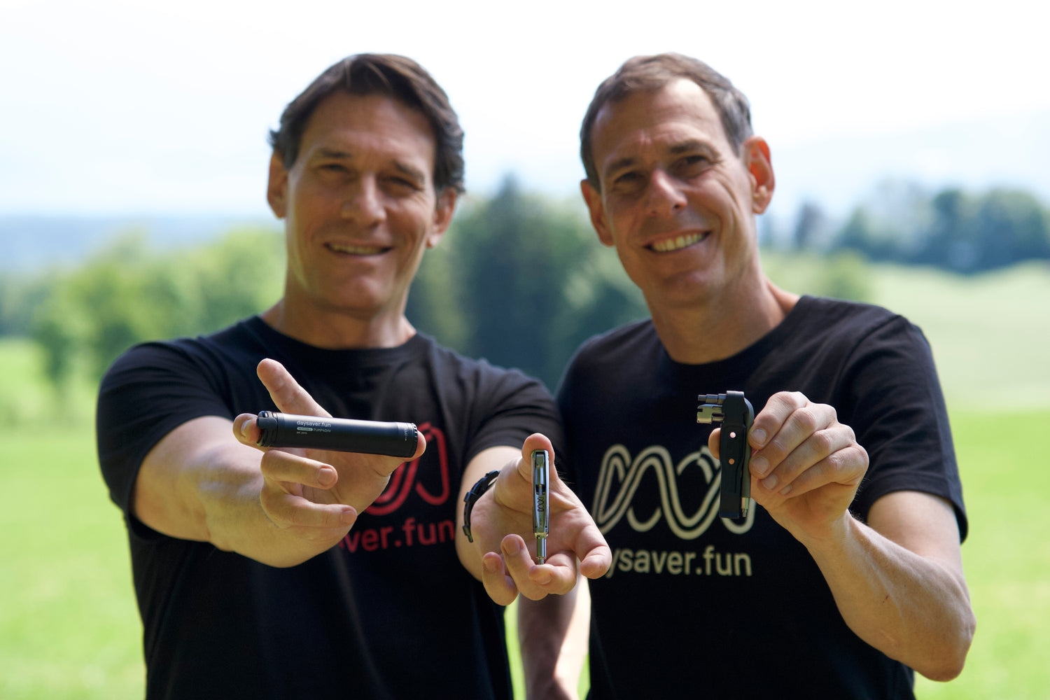 Two men in black t-shirts holding small devices outdoors with a blurred natural background