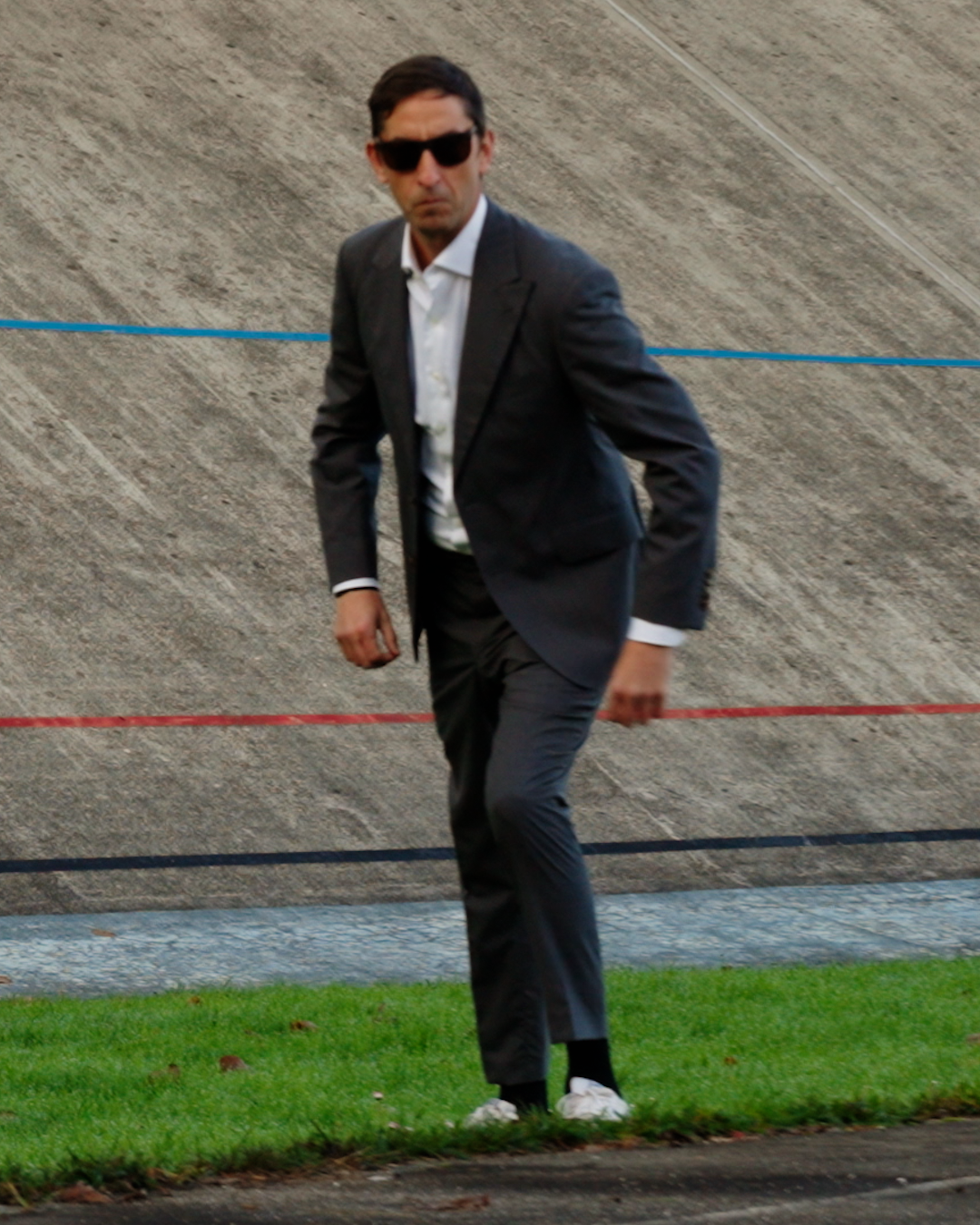 Man in a suit and sunglasses walking on grass with a concrete wall in the background